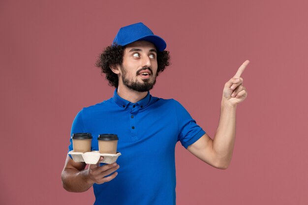 Vista frontale del corriere maschio in protezione uniforme blu con tazze di caffè di consegna sulle sue mani sulla parete rosa