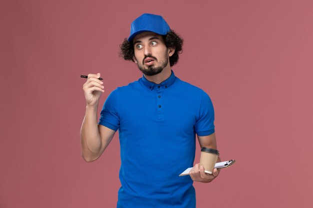 Vista frontale del corriere maschio in uniforme blu e cappuccio con la tazza di caffè di consegna e il blocco note sulle sue mani scrivendo note sul muro rosa