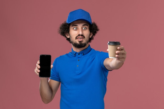 Vista frontale del corriere maschio in uniforme blu e cappuccio con la tazza di caffè di consegna e il telefono sulle mani sul muro rosa