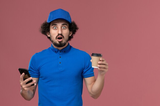 Vista frontale del corriere maschio in uniforme blu e cappuccio con la tazza di caffè di consegna e smartphone sulle sue mani sulla parete rosa