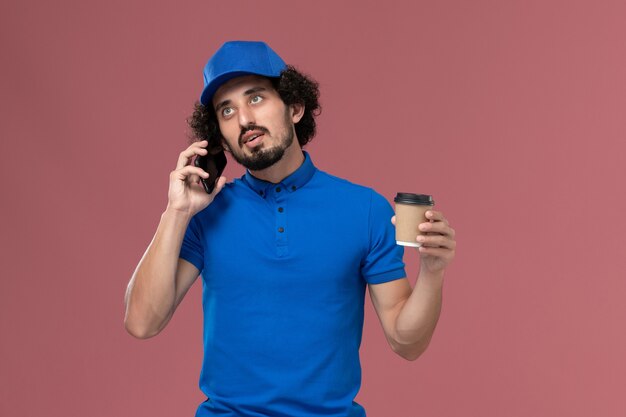 Vista frontale del corriere maschio in uniforme blu e cappuccio con la tazza di caffè di consegna sulle sue mani parlando sul muro rosa