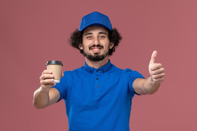 Vista frontale del corriere maschio in uniforme blu e cappuccio con la tazza di caffè di consegna sulle sue mani sul muro rosa