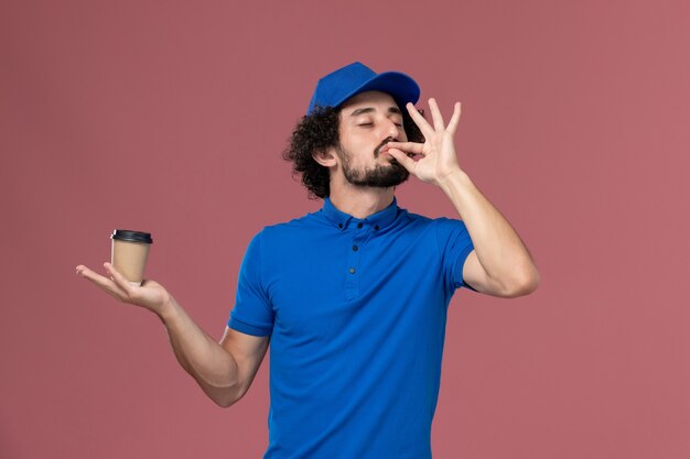 Vista frontale del corriere maschio in uniforme blu e cappuccio con la tazza di caffè di consegna sulle sue mani sul muro rosa