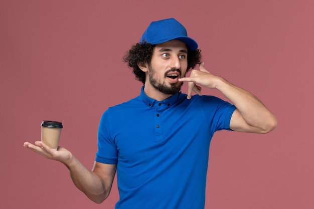 Vista frontale del corriere maschio in uniforme blu e cappuccio con la tazza di caffè di consegna sulle sue mani sul muro rosa