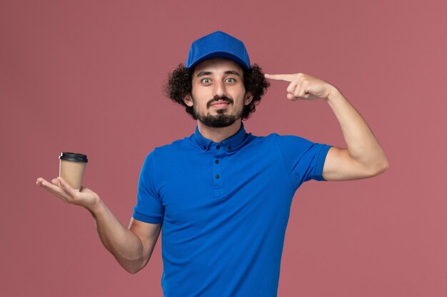 Vista frontale del corriere maschio in uniforme blu e cappuccio con la tazza di caffè di consegna sulle sue mani sul muro rosa