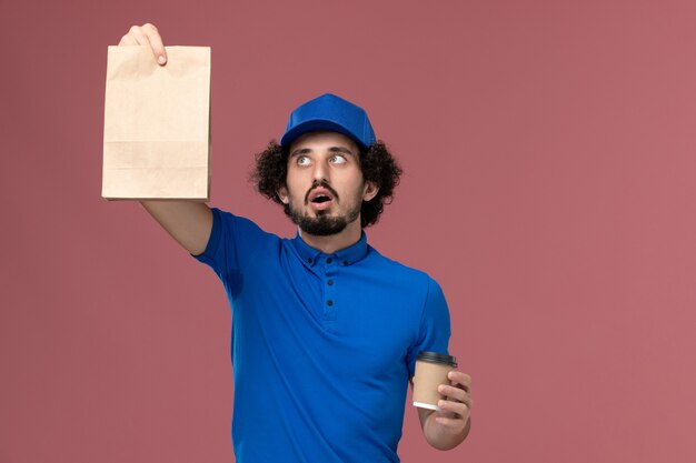 Vista frontale del corriere maschio in uniforme blu e cappuccio con tazza di caffè di consegna e pacchetto di cibo sulle mani sulla parete rosa