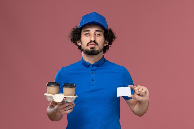 Vista frontale del corriere maschio in uniforme blu e cappuccio con tazze di caffè di consegna e carta sulle sue mani sul muro rosa