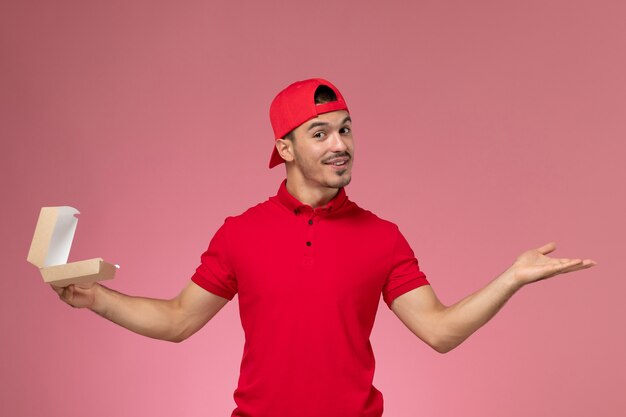 Vista frontale del corriere maschio in uniforme rossa e cappuccio che tiene piccolo pacchetto di consegna sulla parete rosa chiaro