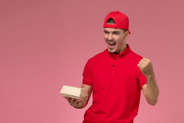 Vista frontale del corriere maschio in uniforme rossa e cappuccio che tiene piccolo pacco di consegna che incoraggia sulla parete rosa
