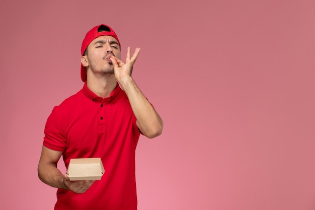 Vista frontale del corriere maschio in uniforme rossa e cappuccio che tiene piccolo pacco di consegna sulla parete rosa