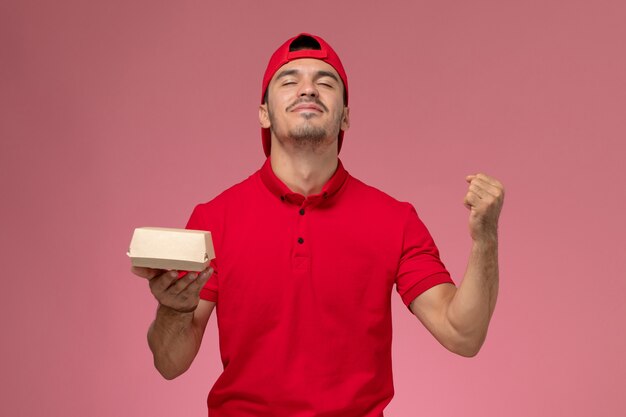 Vista frontale del corriere maschio in uniforme rossa e cappuccio che tiene piccolo pacco di consegna sulla parete rosa