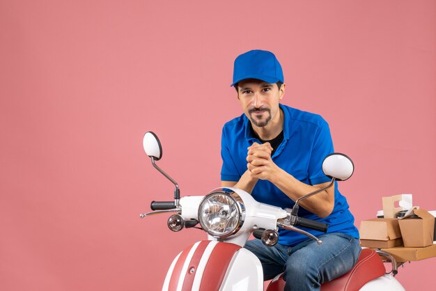 Vista frontale del corriere sorridente che indossa un cappello seduto su uno scooter su sfondo color pesca pastello
