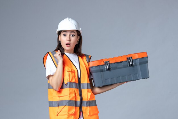 Vista frontale del costruttore femminile che tiene la cassetta degli attrezzi sul muro grigio