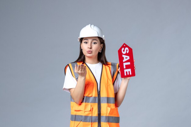 Vista frontale del costruttore femminile che tiene la scheda di vendita rossa sul muro grigio
