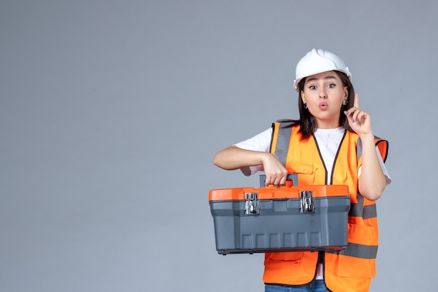 Vista frontale del costruttore femminile con valigetta per attrezzi pesante sul muro grigio