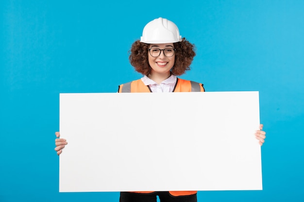 Vista frontale del costruttore femminile in uniforme che tiene lo scrittorio normale bianco sulla parete blu