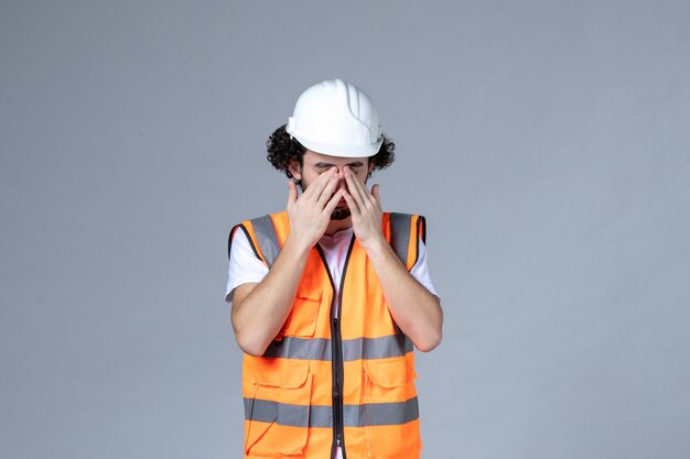 Vista frontale del costruttore maschio che indossa un giubbotto di avvertimento con casco di sicurezza e chiude gli occhi sulla parete grigia dell'onda