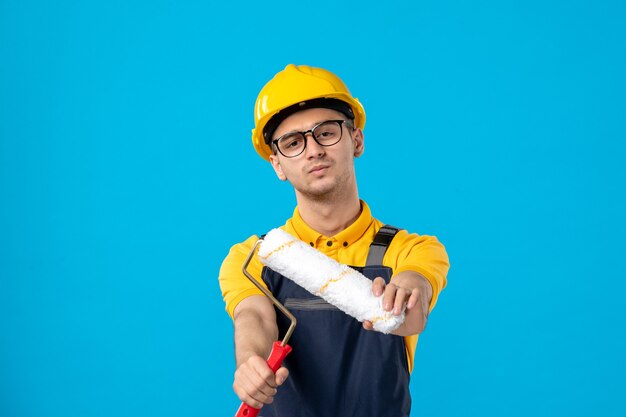 Vista frontale del costruttore maschio in uniforme con rullo di vernice sulla parete blu