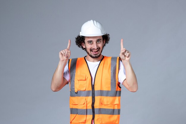 Vista frontale del costruttore maschio sorridente che indossa un giubbotto di avvertimento con casco di sicurezza e punta verso l'alto sulla parete grigia dell'onda