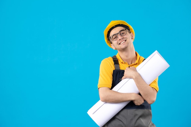 Vista frontale del costruttore maschio sorridente in uniforme gialla con piano di carta sull'azzurro
