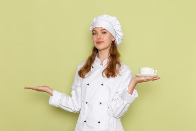 Vista frontale del cuoco femminile in vestito bianco del cuoco che sorride e che tiene tazza bianca con caffè sulla parete verde