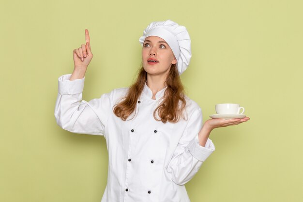 Vista frontale del cuoco femminile in vestito bianco del cuoco che tiene tazza di caffè sulla parete verde chiaro