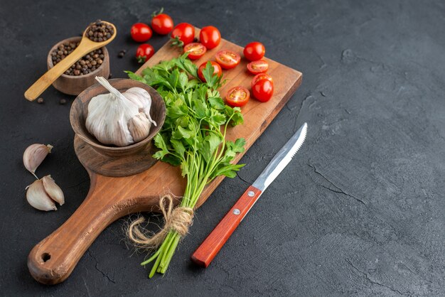 Vista frontale del fascio verde fresco intero di pomodori tagliati aglio su tagliere di legno coltello pepe su superficie nera in difficoltà