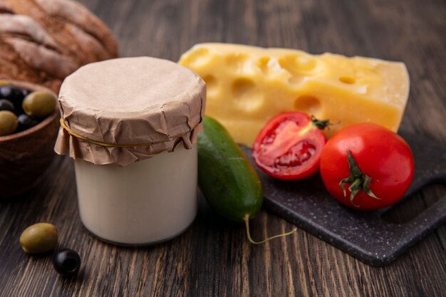 Vista frontale del formaggio maasdam con pomodori e cetrioli su un supporto con olive con yogurt sul tavolo