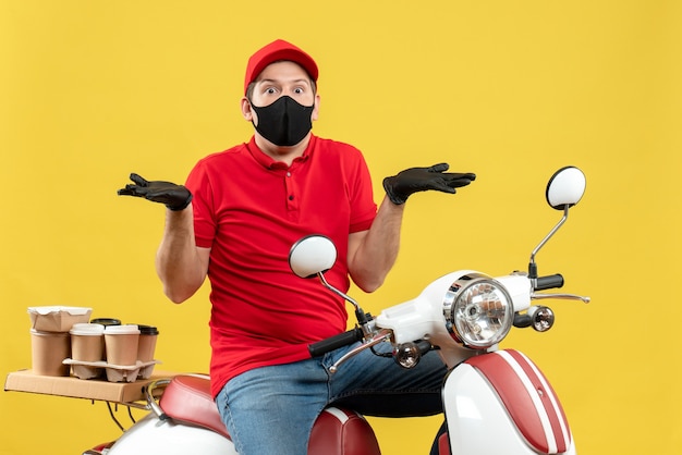 Vista frontale del giovane adulto curioso che indossa la camicetta rossa e guanti del cappello nella mascherina medica che trasporta l'ordine che si siede sullo scooter che indica entrambi i lati su fondo giallo