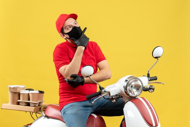 Vista frontale del giovane adulto premuroso che indossa la camicetta rossa e guanti del cappello nella mascherina medica che trasporta l'ordine che si siede sul motorino su fondo giallo