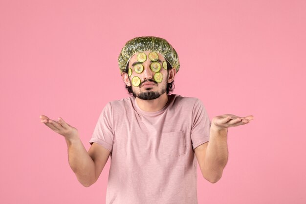 vista frontale del giovane che applica la maschera di cetriolo sul viso sul muro rosa