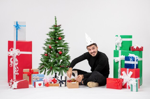 Vista frontale del giovane che si siede intorno ai regali di festa che decorano il piccolo albero sulla parete bianca