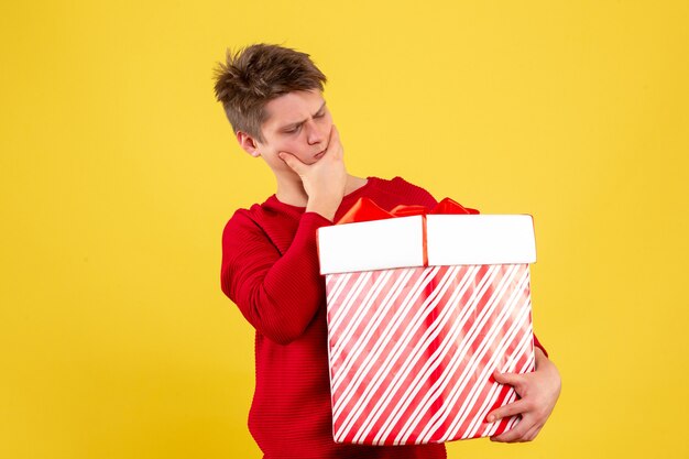 Vista frontale del giovane che tiene grande regalo di Natale pensando sulla parete gialla