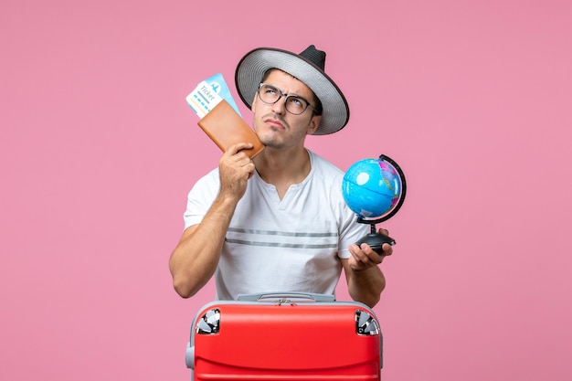 Vista frontale del giovane che tiene i biglietti per le vacanze e un piccolo globo sul muro rosa pink