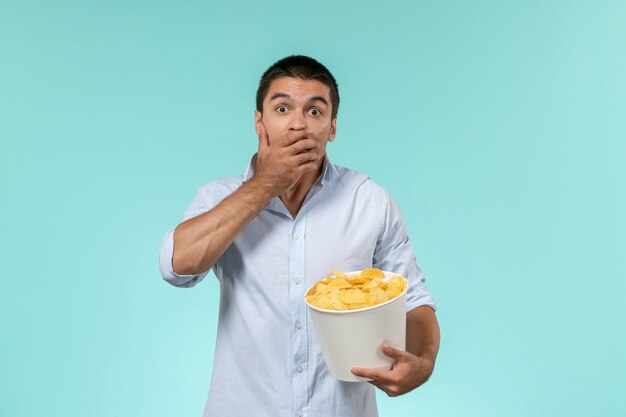 Vista frontale del giovane che tiene il cestino con le patatine fritte che mangia e che guarda film scioccato sul cinema di film a distanza solitario della parete blu