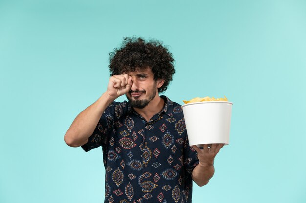 Vista frontale del giovane che tiene il cestino con le patatine fritte e guardare il film che piange sul maschio del teatro di film di film del cinema della parete blu