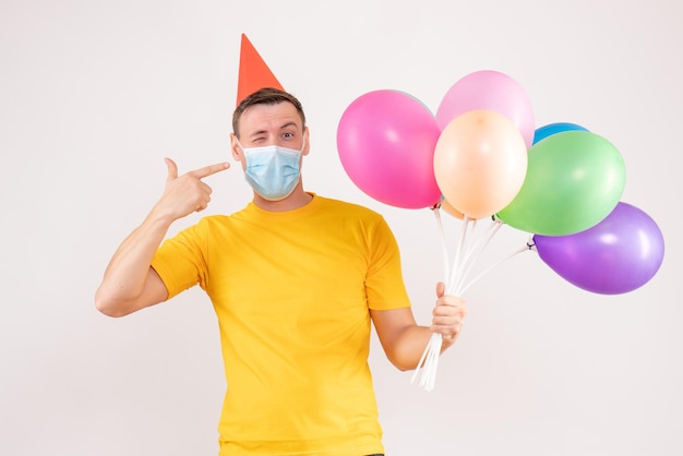 Vista frontale del giovane che tiene palloncini colorati in maschera sul muro bianco