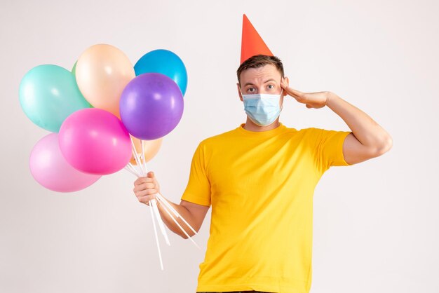 Vista frontale del giovane che tiene palloncini colorati in maschera sul muro bianco