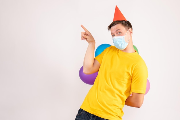 Vista frontale del giovane che tiene palloncini colorati in maschera sul muro bianco