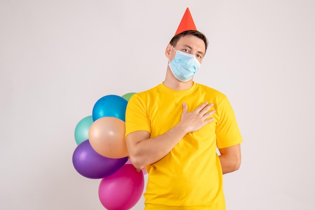 Vista frontale del giovane che tiene palloncini colorati in maschera sul muro bianco