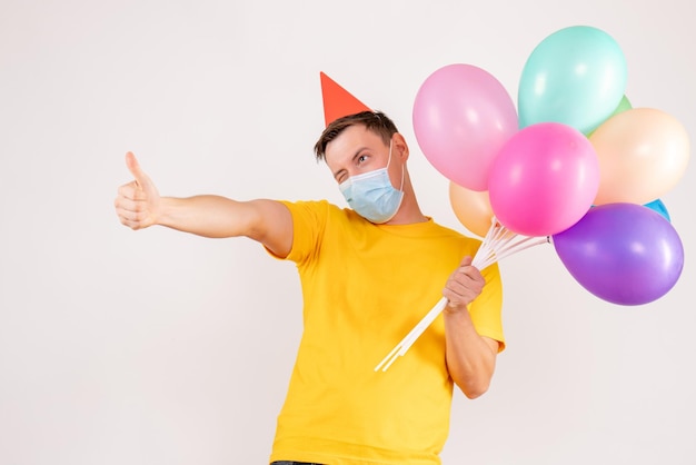 Vista frontale del giovane che tiene palloncini colorati in maschera sul muro bianco