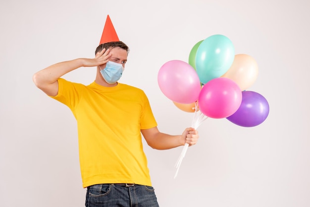 Vista frontale del giovane che tiene palloncini colorati in maschera sul muro bianco