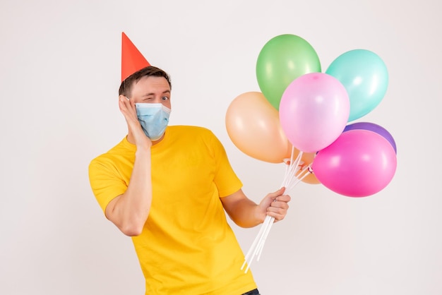 Vista frontale del giovane che tiene palloncini colorati in maschera sul muro bianco