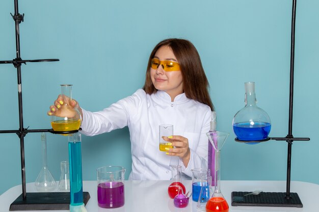 Vista frontale del giovane chimico feman in abito bianco davanti al tavolo di lavoro con soluzioni sulla superficie blu