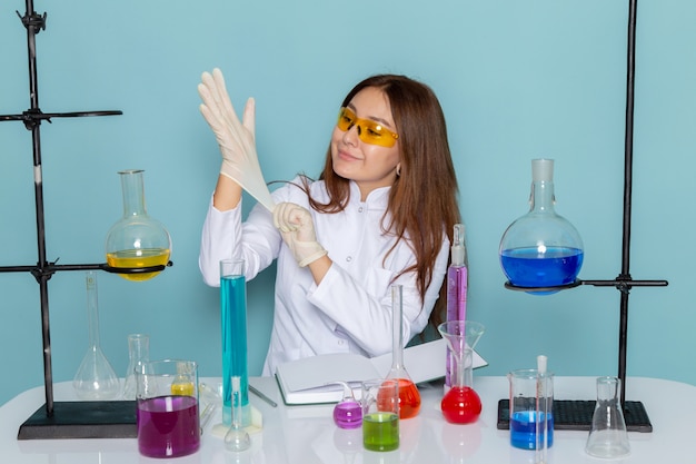 Vista frontale del giovane chimico femmina in abito bianco davanti al tavolo di lavoro con soluzioni e indossando guanti