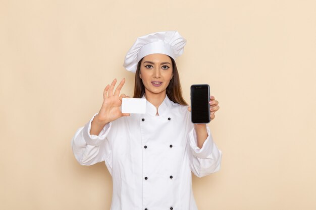 Vista frontale del giovane cuoco femminile in vestito bianco da cuoco che tiene telefono e carta sulla parete bianco-chiaro