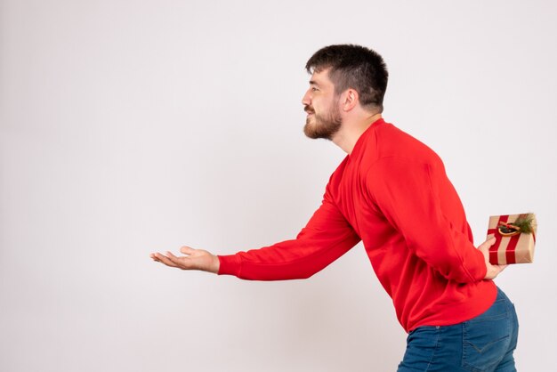 Vista frontale del giovane in camicia rossa che nasconde il regalo di Natale dietro la schiena sul muro bianco