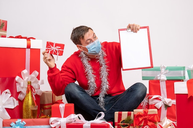 Vista frontale del giovane in maschera seduto intorno ai regali di Natale con nota sul muro bianco