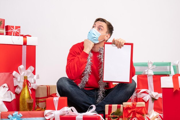 Vista frontale del giovane in maschera seduto intorno ai regali di Natale con nota sul muro bianco