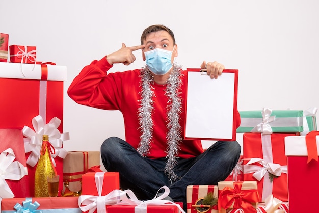 Vista frontale del giovane in maschera seduto intorno ai regali di Natale con nota sul muro bianco